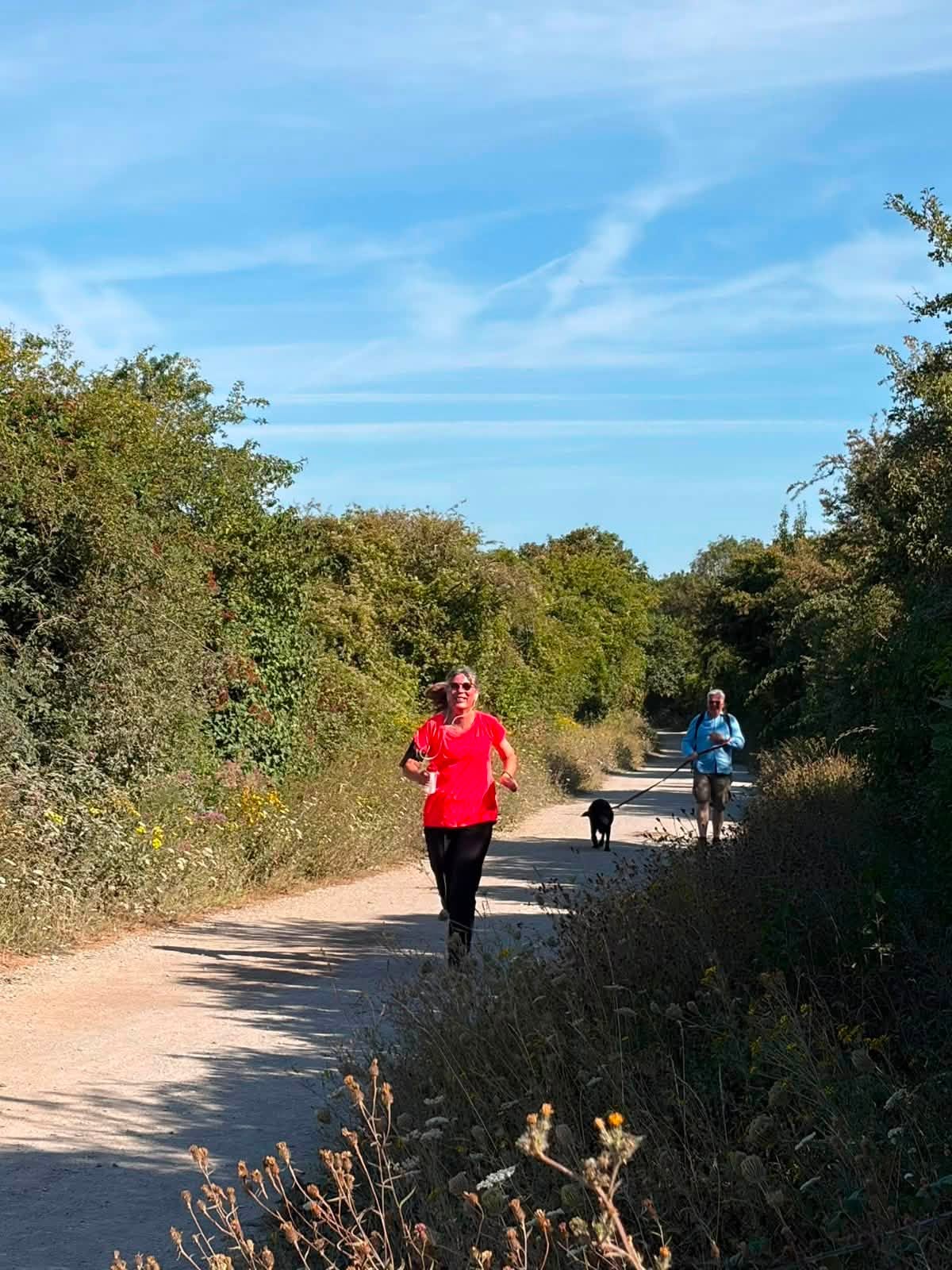 A smiling woman in a bright red top and black leggings jogs along a sandy gravel path lined with green shrubs and wildflowers under a blue sky with wispy clouds. Behind her, a man in a light blue shirt walks a black dog on a long lead.