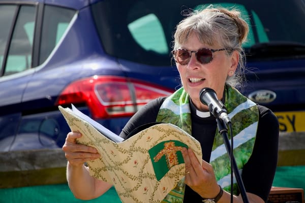 A priest in a green stole reads from a book into a microphone outdoors, with a blue car behind her.
