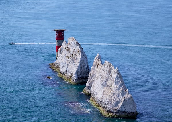The Needles, two chalk stacks and a red-and-white lighthouse, rising from a calm blue sea, with a small boat passing behind.